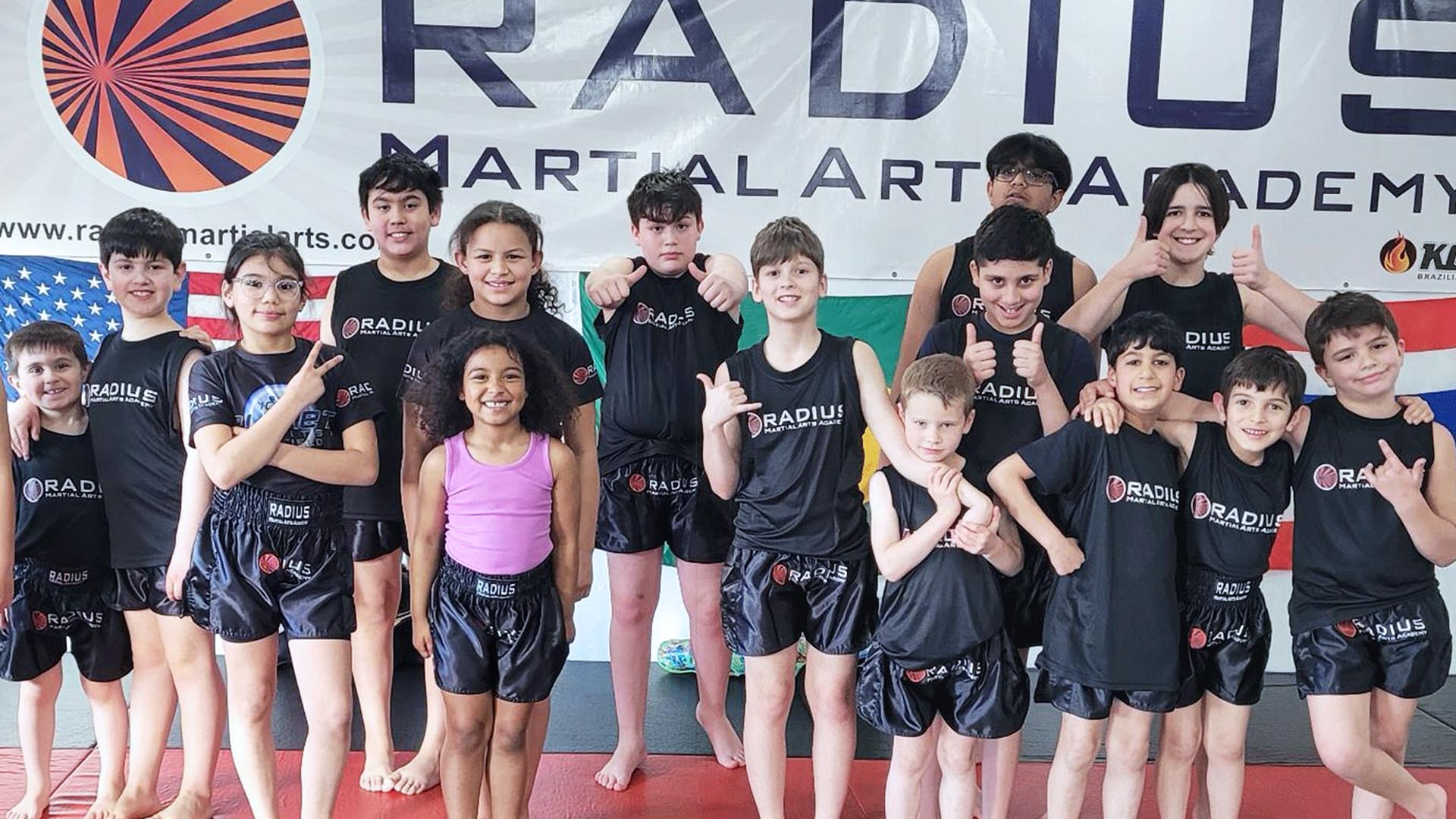 Group of smiling kids in Radius branded shirts and Muay Thai shorts posing beneath the Radius Martial Arts Academy banner in Fairfield, CT