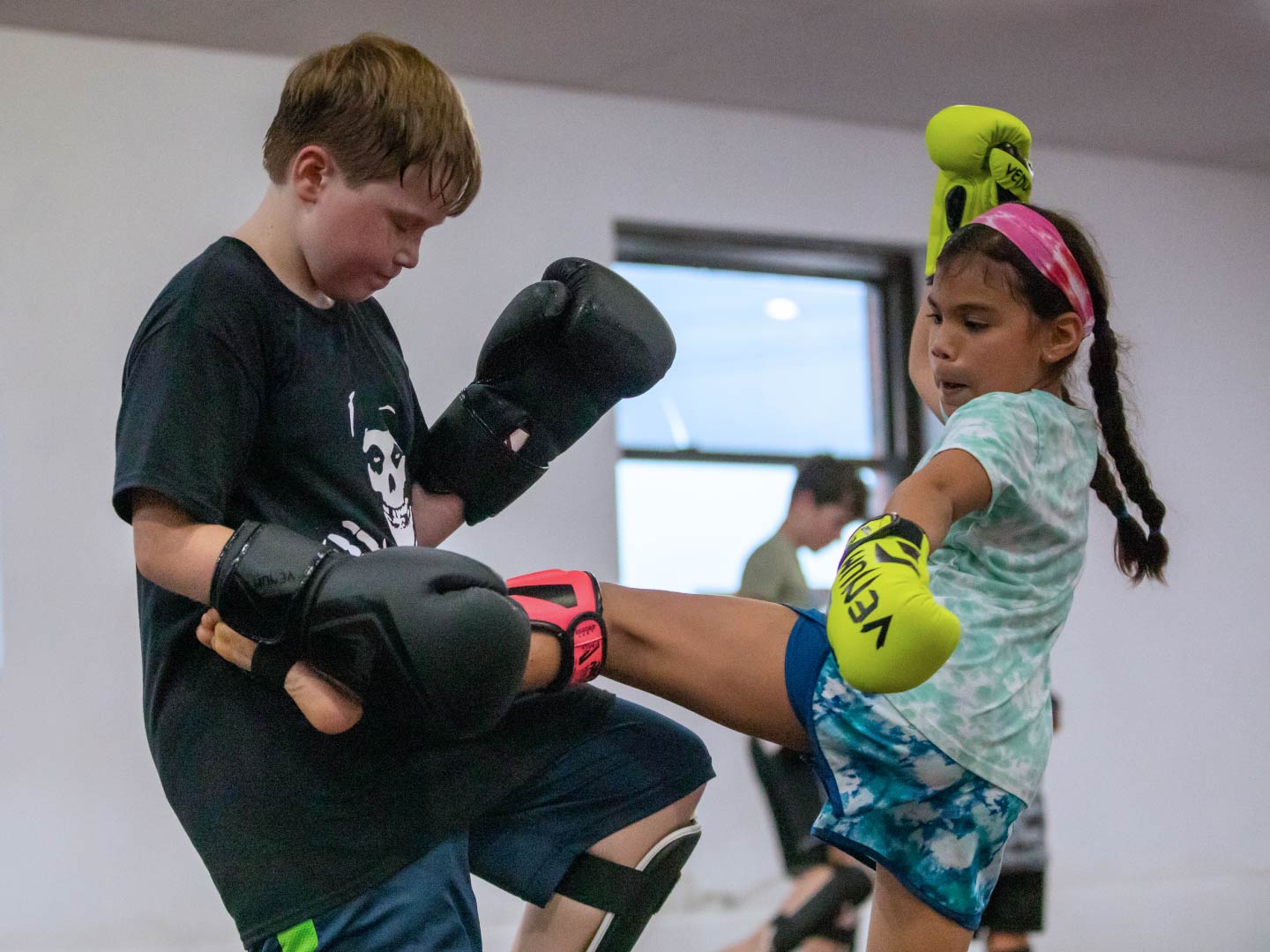 Young girl in boxing gloves throwing a body kick to a partner holding a defensive guard during a kids kickboxing class at Radius Martial Arts in Fairfield, CT