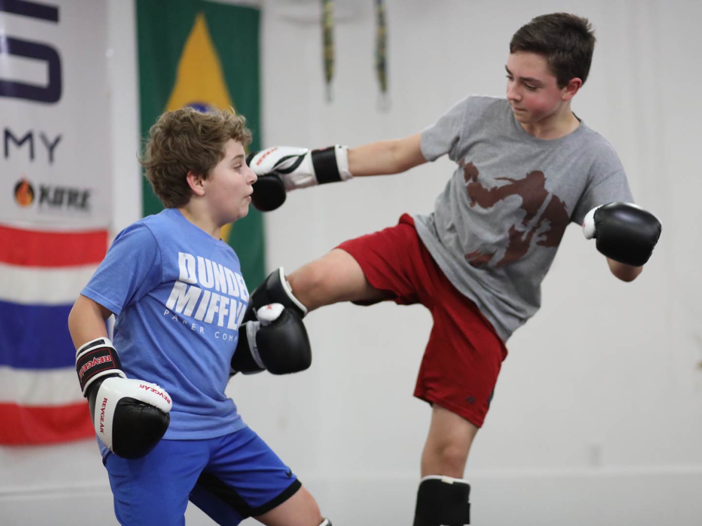 Two boys in boxing gloves and shin guards practicing kickboxing, one throwing a front kick, during a kids class at Radius Martial Arts in Fairfield, CT